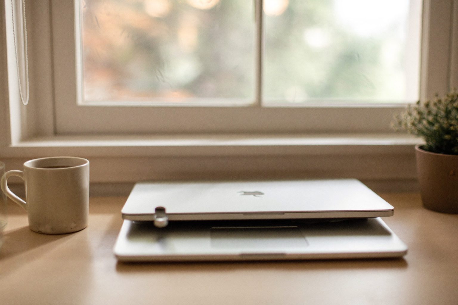 Een opgeruimd bureau bij het raam met een dichtgeklapte laptop, een kop koffie en een plant