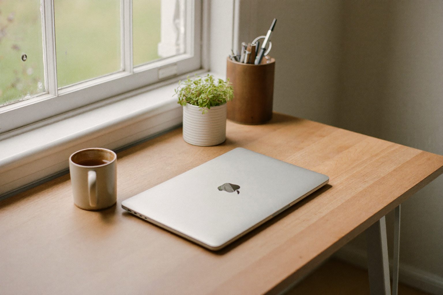 Een opgeruimd bureau bij het raam met een dichtgeklapte laptop, een kop koffie en een plant