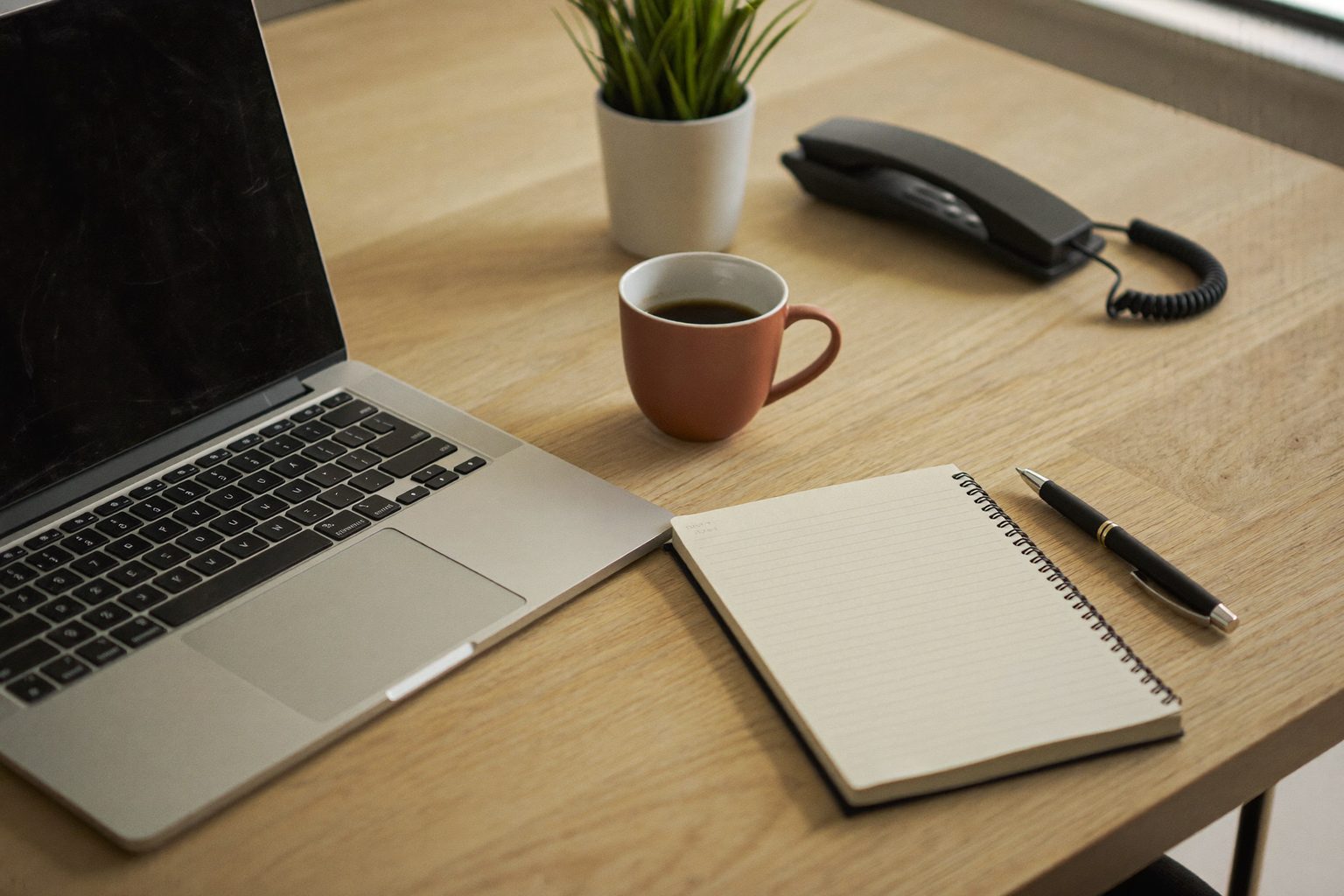 Een opgeruimde, lichte kantoorwerkplek met laptop, notitieblok, koffie en een bureautelefoon